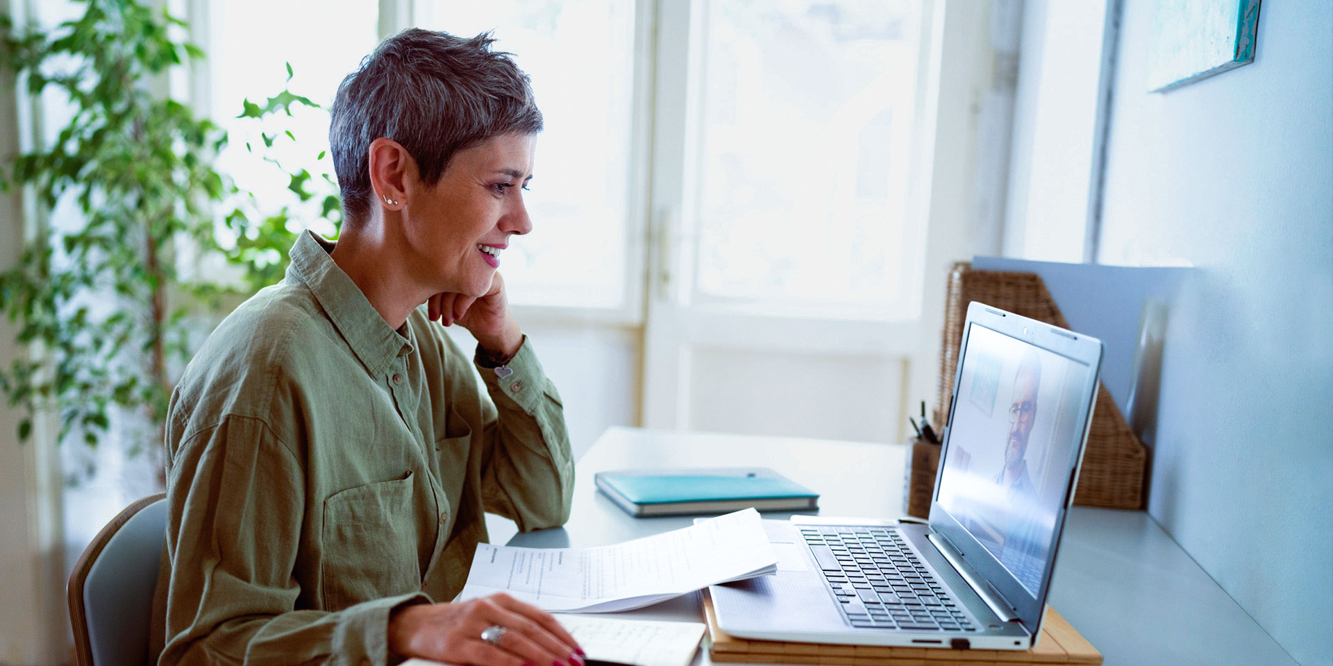 A woman with short grey hair smiling while participating in a laptop video call at her desk.