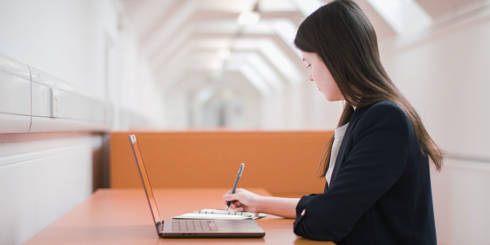 A young woman sitting at a long desk with a open laptop, writing notes in a notebook.