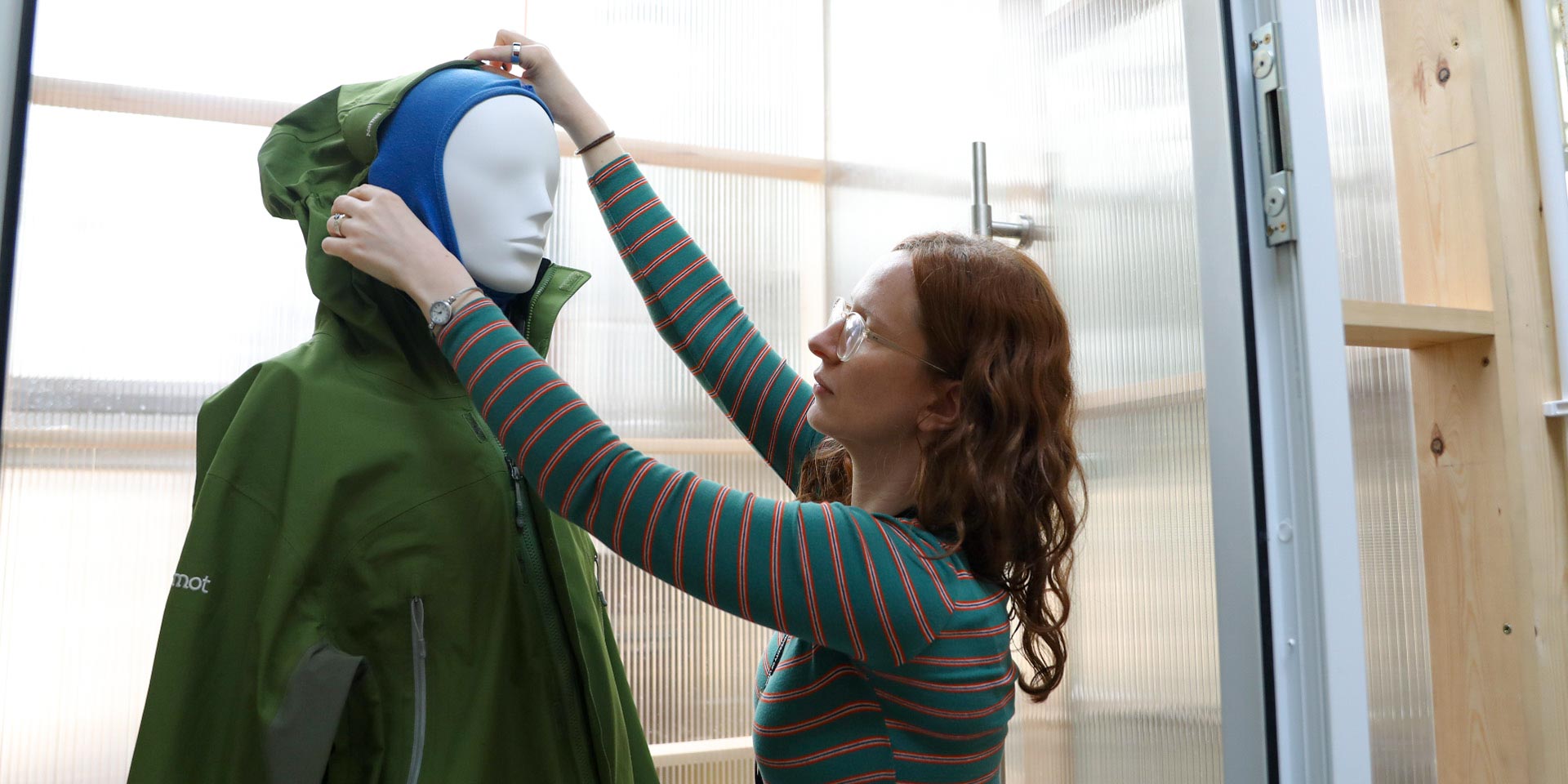 A woman, Alice Hazlehurst, adjusting a green waterproof hooded jacket on a mannequin inside a testing room