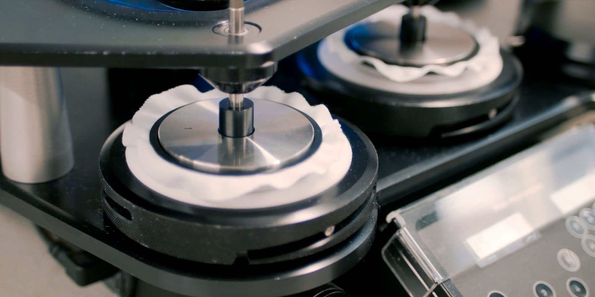 Close-up of a textile testing machine with circular metal heads holding white fabric swatches on a testing station.